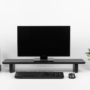 May include: A black computer monitor sits on a black wooden monitor stand. A black keyboard and mouse are on a white desk in front of the monitor. A small potted plant is on the right side of the desk.