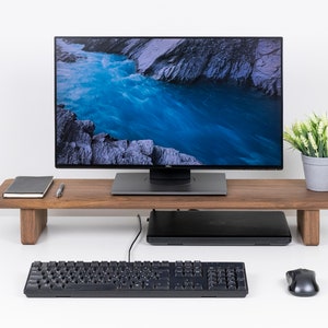 Puede incluir: Un soporte de monitor de madera con un monitor negro, un port&aacute;til negro, un teclado negro, un rat&oacute;n negro y una planta en maceta con hojas verdes sobre una superficie blanca.