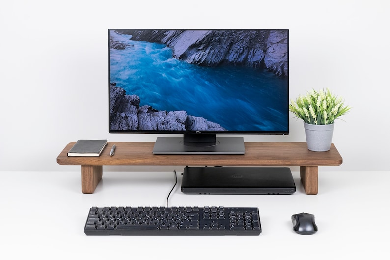 Puede incluir: Un soporte de monitor de madera con un monitor negro, un port&aacute;til negro, un teclado negro, un rat&oacute;n negro y una planta en maceta con hojas verdes sobre una superficie blanca.