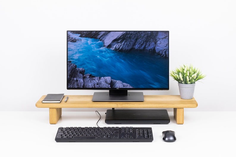 May include: A wooden monitor stand with a black monitor, keyboard, and mouse on a white desk. The monitor is displaying a scenic image of a river flowing through a rocky landscape.