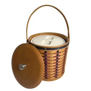 May include: Woven basket with a wooden handle and lid. The basket features a cream-colored interior lid and a woven design with red and blue accents. The wooden lid has a silver knob. The basket is designed for carrying items.