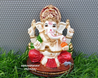 May include: A white and gold Ganesha statue with red and green clothing, sitting on a brown base. The statue is on a bed of green grass.