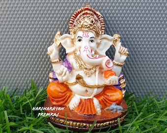 May include: A white Ganesha figurine with a red, gold and purple headdress, sitting on a brown base with a gold trim. The figurine is holding a small elephant in its right hand and a conch shell in its left hand. The figurine is surrounded by green grass.