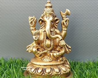 Puede incluir: Una estatua de metal dorado de la deidad hindú Ganesha. La estatua representa a Ganesha sentado con la trompa levantada y sosteniendo un pequeño hacha en su mano superior derecha. La estatua está sobre una base circular con detalles decorativos.