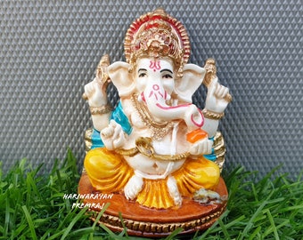 May include: A small, white, seated statue of Ganesha, the Hindu god of wisdom and prosperity. The statue is decorated with gold, red, and blue paint. Ganesha is holding a small, orange object in his right hand. The statue is sitting on a brown base with gold trim. The statue is on a bed of green grass.