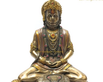 Może przedstawiać: Złota statua hinduskiego boga Hanuman, siedzącego w medytacyjnej pozie. Ma na sobie koronę, biżuterię i czerwoną i złotą szatę. Siedzi na złotej platformie z książką i różańcem.