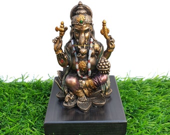 Puede incluir: Una estatua dorada y de bronce de Ganesha, el dios hindú de la sabiduría y la prosperidad, sentado en una flor de loto. La estatua está sobre una base de madera negra y está rodeada de hierba verde.