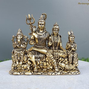 May include: A gold-colored metal statue of the Hindu deities Shiva, Parvati, and Ganesha. The statue depicts the three deities sitting together, with Shiva holding a trident and Parvati holding a lotus flower. Ganesha is sitting on a throne with his trunk raised.