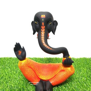May include: Black and orange Hindu deity Ganesha statue with a raised hand and a curled trunk. The statue is sitting on a green grassy surface.