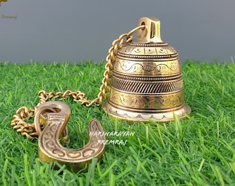 Può includere: Una campana in ottone dorata con intricati disegni incisi è appesa a una catena con un gancio. La campana è su una superficie di erba artificiale verde.
