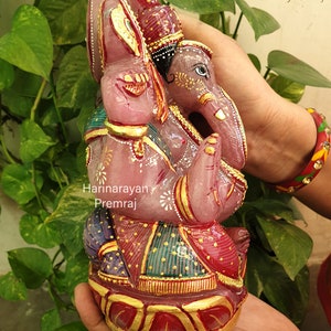 May include: A pink, hand-carved, gemstone Ganesha statue with gold accents and intricate details. The statue is sitting on a lotus base and is adorned with colorful clothing.