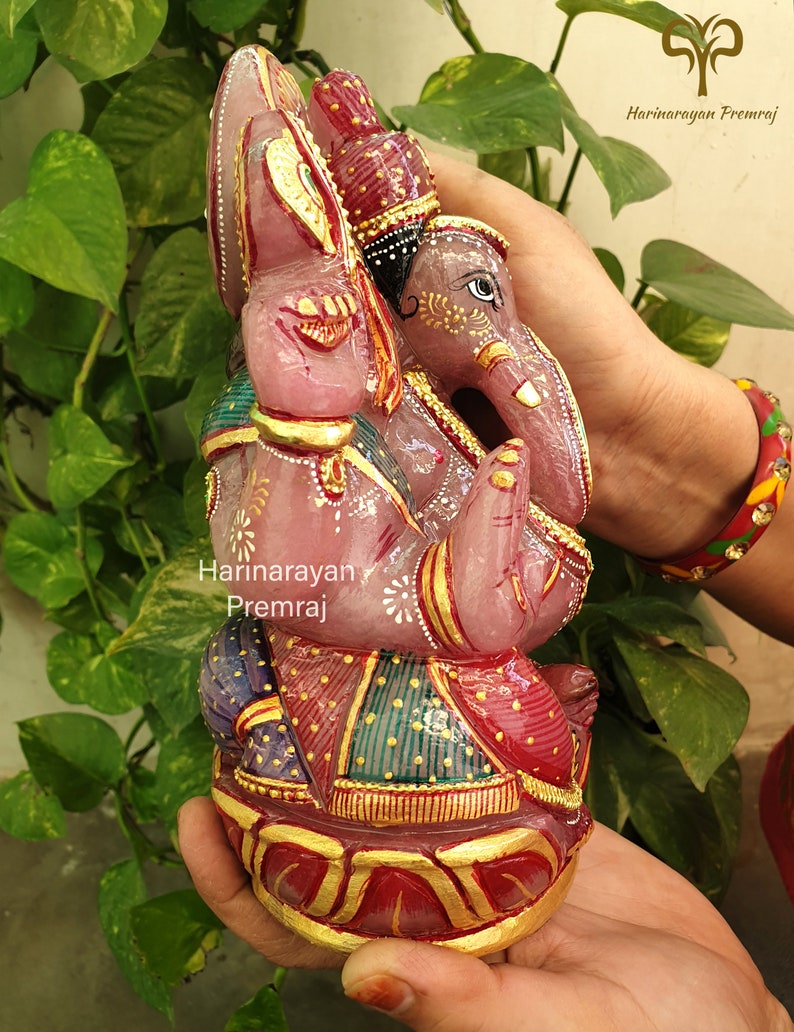 May include: A pink, hand-carved, gemstone Ganesha statue with gold accents and intricate details. The statue is sitting on a lotus base and is adorned with colorful clothing.