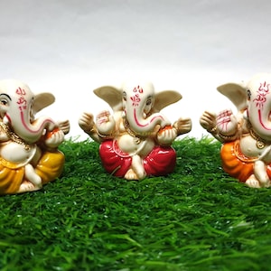 May include: Three small white ceramic statues of the Hindu deity Ganesha. Each statue is sitting with its legs crossed and has a red, yellow, or orange dhoti. The statues are on a green artificial grass surface.