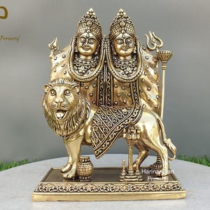 Puede incluir: Estatua de metal dorado de dos diosas sentadas sobre un león. Las diosas llevan ropa y joyas adornadas. El león está de pie sobre una base con detalles intrincados.