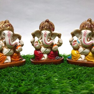 May include: Three colorful Ganesha figurines, each with a different colored robe, sitting on a brown base. The figurines are made of white painted resin and have gold accents. The figurines are sitting on a green grass surface.