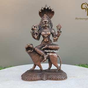 May include: Bronze-colored statue of a deity riding a lion. The deity has multiple arms and wears a crown with cobra heads. The lion is detailed with etched lines. The statue is on a decorative base. The text "Harinarayan Premraj" is visible.