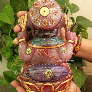 May include: A pink and purple Ganesha statue with gold accents and intricate details. The statue is made of stone and is sitting on a lotus base.
