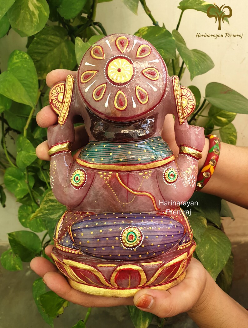 May include: A pink and purple Ganesha statue with gold accents and intricate details. The statue is made of stone and is sitting on a lotus base.