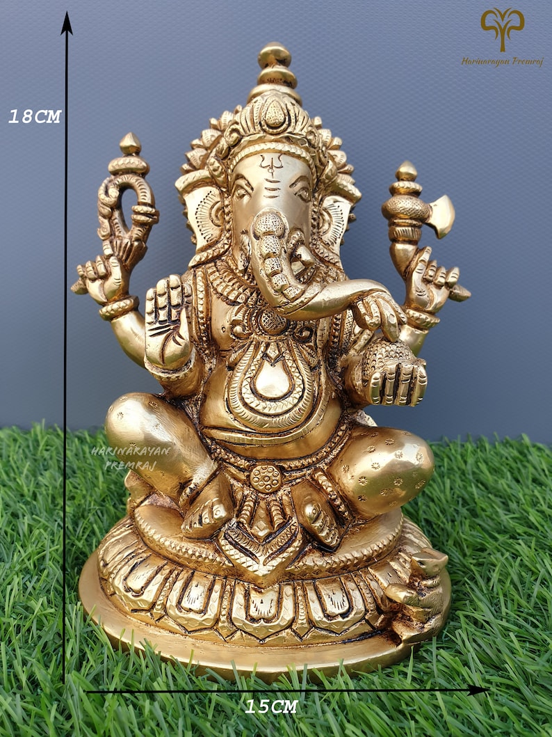 Puede incluir: Una estatua de bronce dorado de Ganesha, el dios hind&uacute; de la sabidur&iacute;a y la prosperidad, sentado sobre una base de loto. La estatua mide 18 cm de alto y 15 cm de ancho.