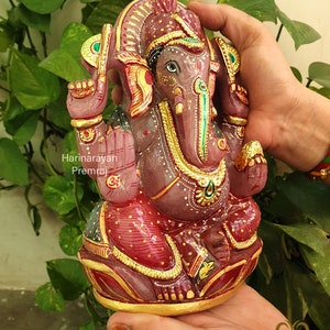 May include: A pink and gold Ganesha statue with intricate details. The statue is made of stone and is decorated with gold paint and jewels. The statue is sitting on a lotus flower base.