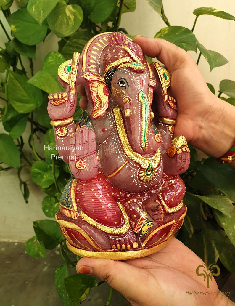 May include: A pink and gold Ganesha statue with intricate details. The statue is made of stone and is decorated with gold paint and jewels. The statue is sitting on a lotus flower base.