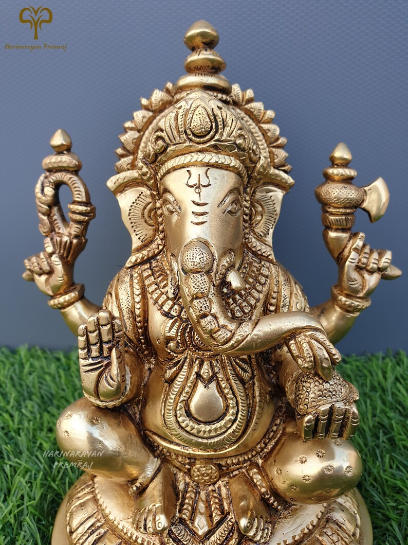 Puede incluir: Una estatua de metal dorada de Ganesha, el dios hind&uacute; de la sabidur&iacute;a y la prosperidad. La estatua est&aacute; intrincadamente detallada y presenta a Ganesha sentado en un trono de loto con su trompa levantada y sosteniendo un dulce modaka en su mano.