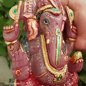 May include: A pink, hand-carved Ganesha statue with gold and green accents. The statue is sitting on a red base with gold and white accents. The statue is made of rose quartz.