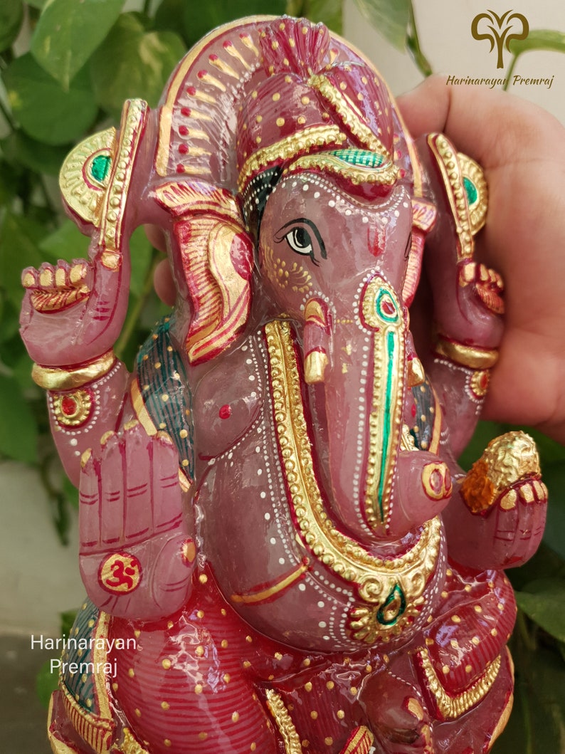 May include: A pink, hand-carved Ganesha statue with gold and green accents. The statue is sitting on a red base with gold and white accents. The statue is made of rose quartz.