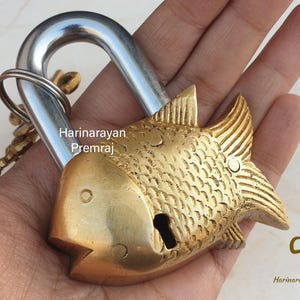 Puede incluir: Un candado de latón con forma de pez con un grillete y una cerradura plateados. El candado tiene un diseño detallado con escamas y aletas.