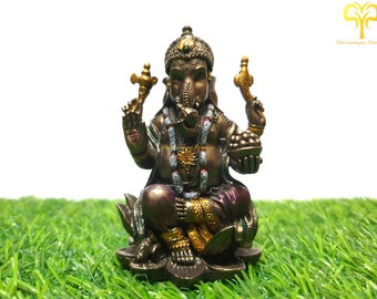 Puede incluir: Una estatua de Ganesha dorada y marrón sentada sobre una base de flor de loto. La estatua sostiene un cuenco de dulces en una mano y un colmillo roto en la otra. La estatua está sentada sobre una superficie de hierba verde.