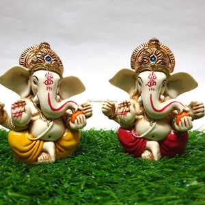May include: Two small, white, decorative Ganesha figurines with gold accents. The figurines are sitting on a green, artificial grass surface. One figurine is holding a yellow fruit and the other is holding an orange fruit. The figurines are wearing yellow and red dhotis.