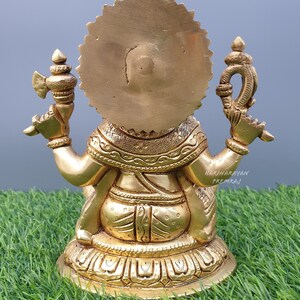 Puede incluir: Una estatua de metal dorado de una deidad, sentada sobre una base con detalles intrincados. La estatua tiene un halo circular detr&aacute;s de la cabeza y sostiene dos objetos en sus manos.