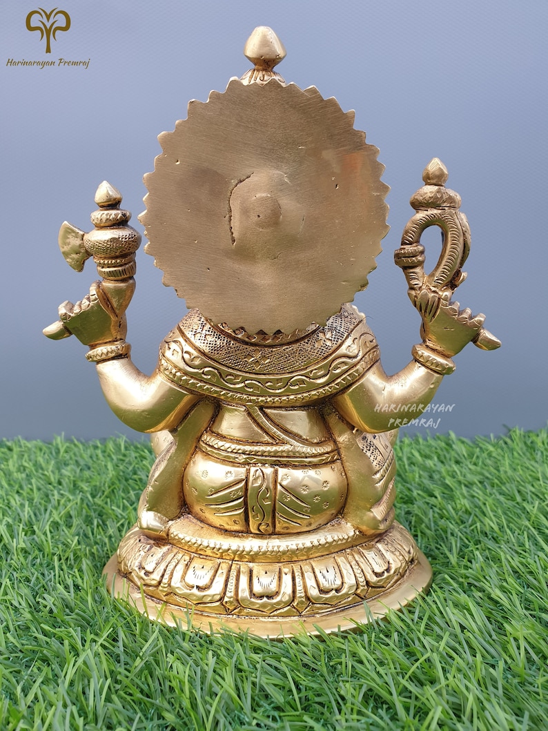 Puede incluir: Una estatua de metal dorado de una deidad, sentada sobre una base con detalles intrincados. La estatua tiene un halo circular detr&aacute;s de la cabeza y sostiene dos objetos en sus manos.