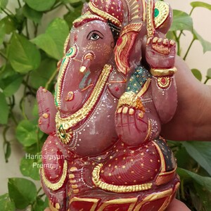 May include: A pink and red hand-painted Ganesha statue with gold accents. The statue is sitting on a lotus base and has a traditional Indian design.