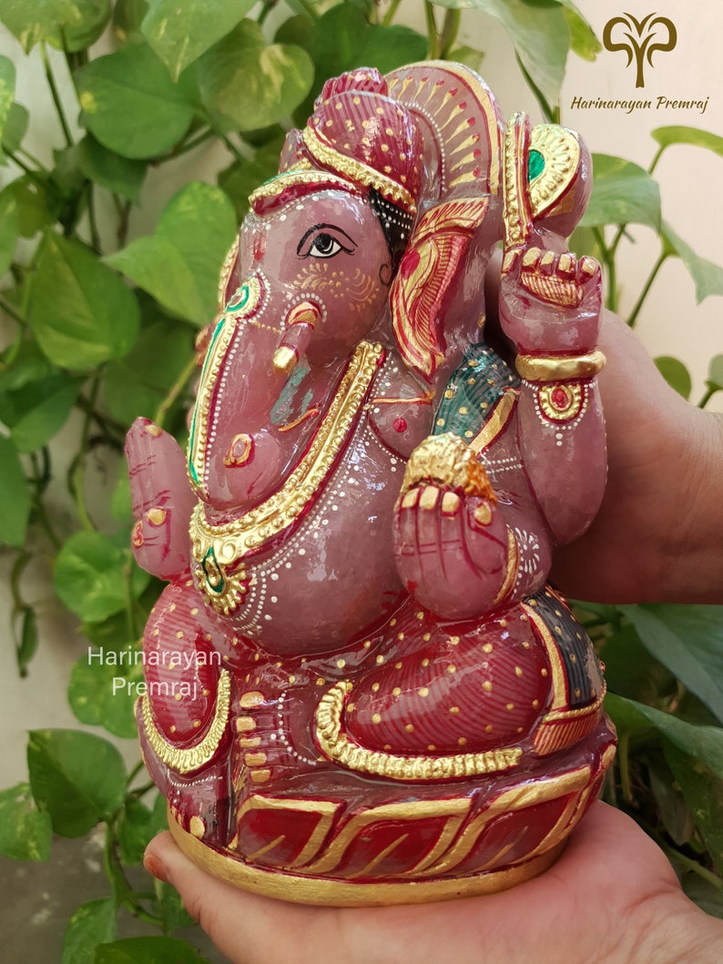 May include: A pink and red hand-painted Ganesha statue with gold accents. The statue is sitting on a lotus base and has a traditional Indian design.
