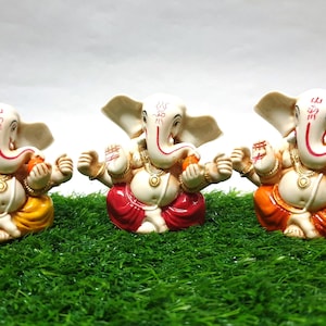 May include: Three white Ganesha figurines with red, orange, and yellow dhotis. Each figurine has a red trunk, gold jewelry, and a red and gold patterned design.