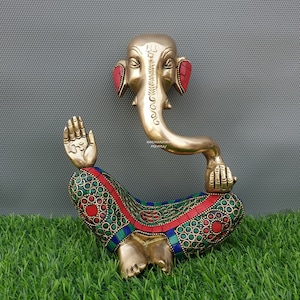May include: A gold-colored brass statue of Ganesha, the Hindu god of wisdom and prosperity, sitting in a meditative pose. The statue is decorated with colorful gemstones and has a red gemstone on each ear.