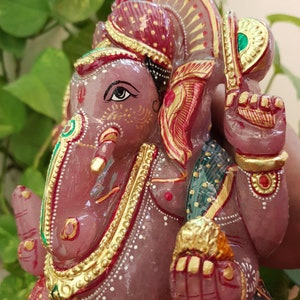 May include: A pink stone sculpture of Ganesha, the Hindu god of beginnings, with gold accents and a red and green patterned cloth. The sculpture is intricately detailed and features a red and gold headdress.