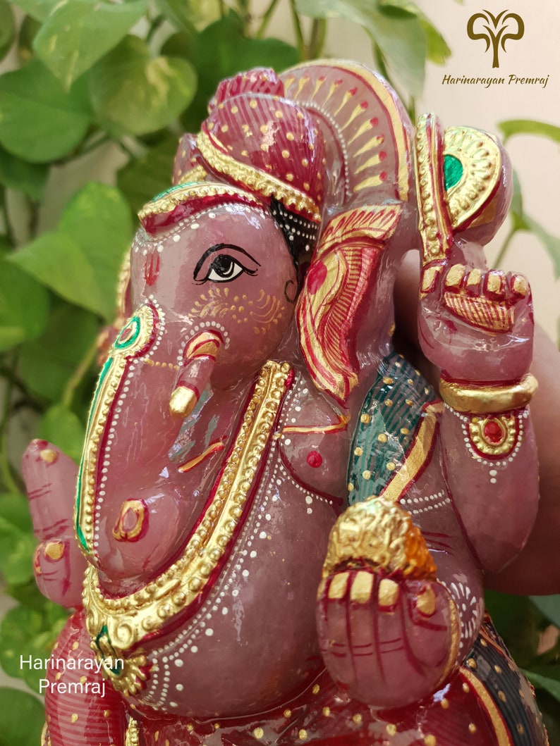 May include: A pink stone sculpture of Ganesha, the Hindu god of beginnings, with gold accents and a red and green patterned cloth. The sculpture is intricately detailed and features a red and gold headdress.