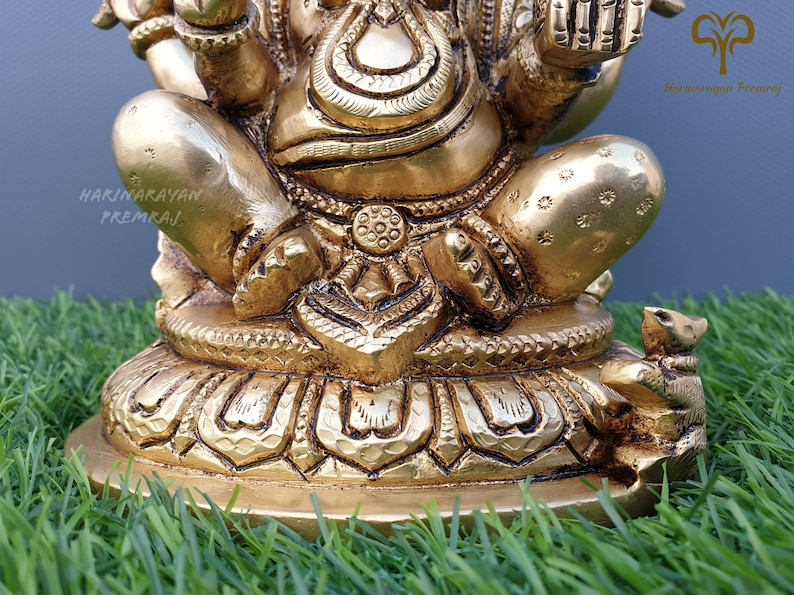 Puede incluir: Una estatua de bronce dorada de Ganesha, el dios hind&uacute; de la sabidur&iacute;a y la prosperidad, sentado sobre una base de loto. La estatua est&aacute; intrincadamente detallada y presenta a Ganesha con su trompa levantada y sosteniendo un dulce en su mano. Un peque&ntilde;o rat&oacute;n est&aacute; sentado en la base de la estatua.