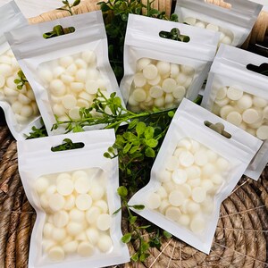 May include: Multiple small white wax melts are packaged in clear plastic bags with white zipper closures. The bags are arranged in a woven basket with green leafy plants.