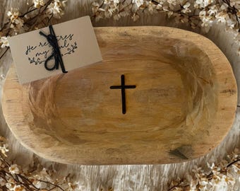 May include: A light brown, oval-shaped wooden bowl with a black cross carved into the center. A small card with the text "He refreshes my soul" is tied with black twine. White flowers surround the bowl.