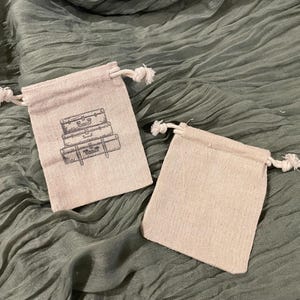May include: Two small, beige drawstring bags made of a textured fabric. One bag features a black illustration of stacked vintage suitcases. The bags are on a green, textured surface.
