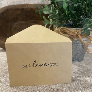 May include: A light brown kraft paper envelope with a triangular flap. The front of the envelope features the text p.s. i love you printed in a black, handwritten-style font centered on the lower portion of the paper surface.