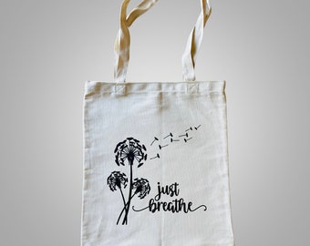 May include: A white canvas tote bag with black text that reads "just breathe" and a black silhouette of three dandelions with seeds blowing in the wind.