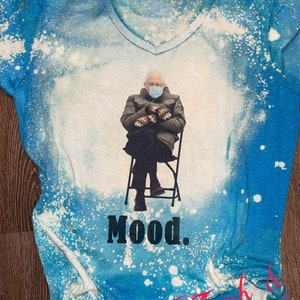 May include: A blue t-shirt with a white bleach design. The design features a person wearing a face mask sitting in a chair with the text "Mood." printed below.
