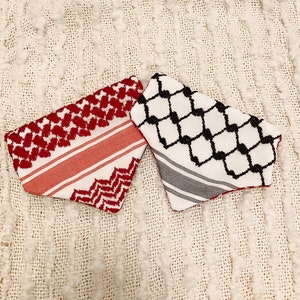 May include: Two keffiyeh scarves. One is red and white with a geometric pattern and stripes. The other is white with a black geometric pattern and gray stripes. Both are folded and displayed on a textured, off-white surface.