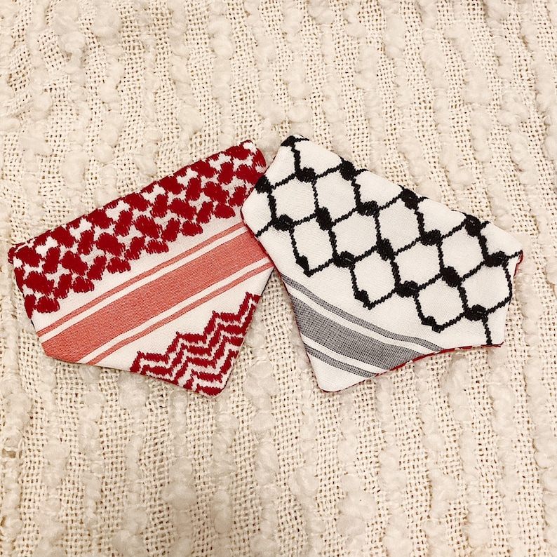 May include: Two keffiyeh scarves. One is red and white with a geometric pattern and stripes. The other is white with a black geometric pattern and gray stripes. Both are folded and displayed on a textured, off-white surface.