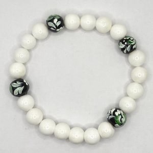 May include: White beaded bracelet with four black beads featuring a green and white floral design. The bracelet is made of round, smooth white beads. The bracelet is a piece of jewelry.