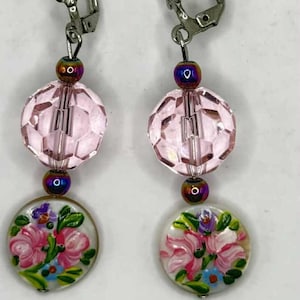 May include: A pair of dangle earrings featuring a floral design. Each earring has a faceted pink bead, a smaller iridescent bead, and a round, hand-painted floral pendant with pink and blue flowers.